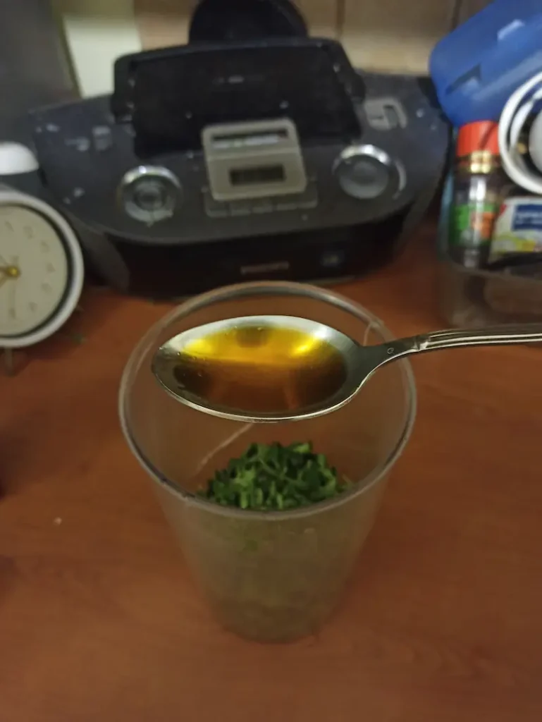 a tablespoon of Calabrian chili oil is being poured over fresh oregano leaves and garlic chops in an immersion food processor